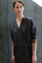 Load image into Gallery viewer, A woman with short brown hair stands before a textured gray backdrop, wearing the Reversible Wrap Dress with Sleeves | Xiang Yun Silk Brown—a dark, long-sleeved wrap dress featuring light brown cuffs. She gazes slightly to her left with a natural expression.