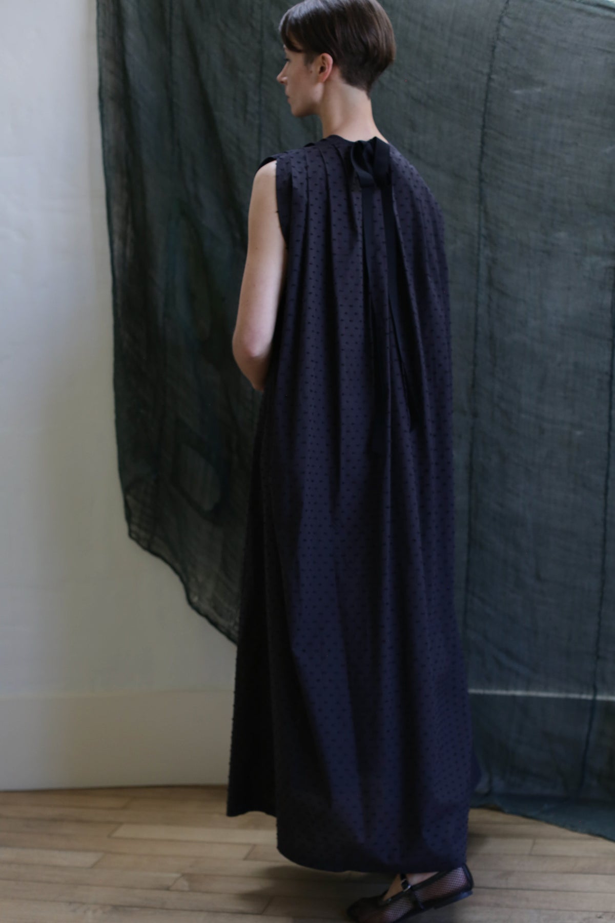 A person with short hair faces away, wearing the Shoulder Pleat Dress | Clip Dot Black with a high gathered collar and shoulder pleats. A sheer dark fabric hangs in the background, above a wooden floor.