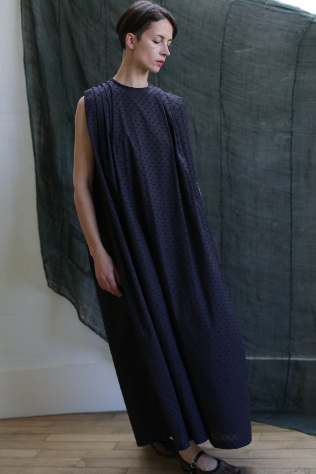 A woman with short brown hair stands indoors against a draped dark green backdrop, wearing the Shoulder Pleat Dress in Clip Dot Black—a sleeveless, floor-length style with subtle shoulder pleats.