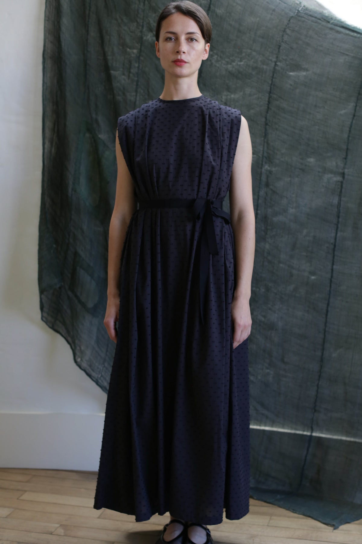 A woman poses indoors in the Shoulder Pleat Dress | Clip Dot Black, featuring a tied waist and pleated shoulders, against a textured dark green fabric backdrop with a wooden floor complementing her neutral expression.