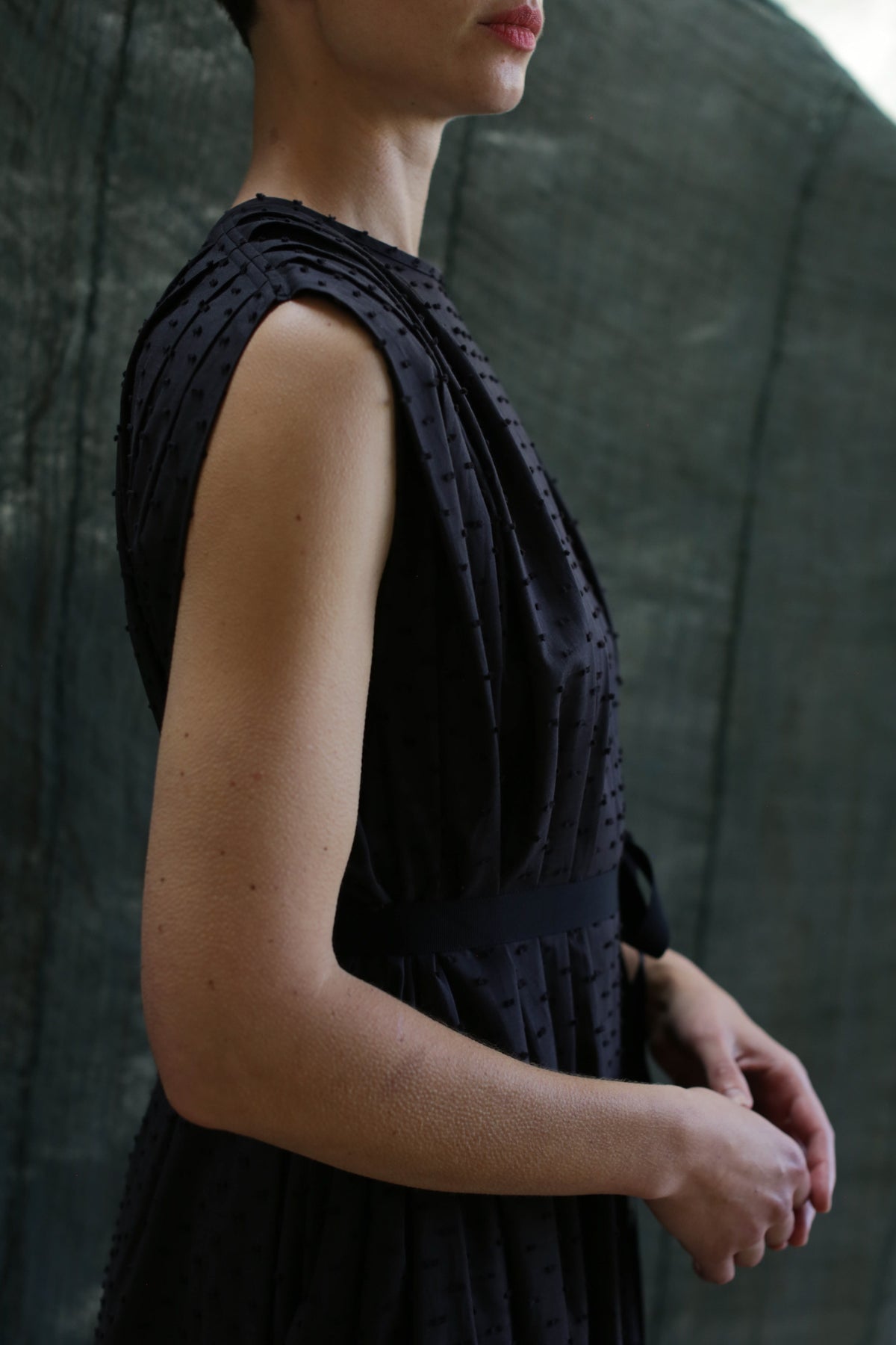 A woman in the Shoulder Pleat Dress | Clip Dot Black stands sideways against a softly-lit dark green background, her face partly visible and hands gently clasped.