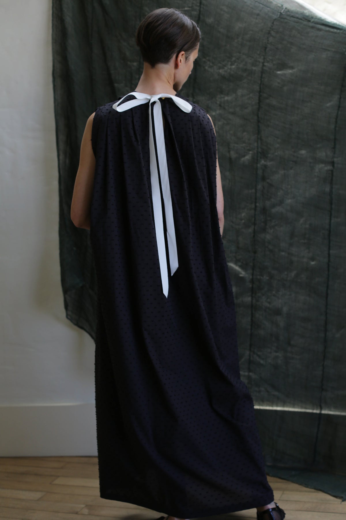 A person with short dark hair stands facing away, wearing the Shoulder Pleat Dress in Clip Dot Black—a long, sleeveless black gown with a white neck ribbon. The textured black clip dot fabric hangs in the background against a white wall and wood floor.