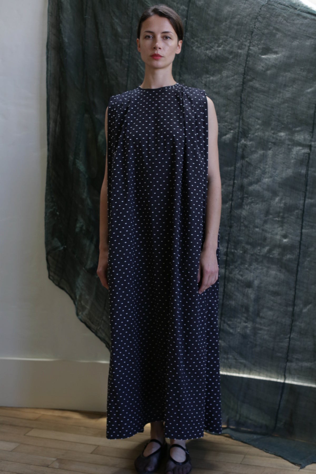 A woman poses indoors on a wooden floor, wearing the Shoulder Pleat Dress in Clip Dot Black & White by Atelier Lan Jaenicke—a long, sleeveless black dress with white dots. Her hair is pulled back and she stands before draped fabric.