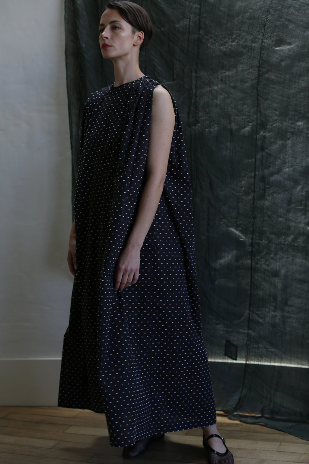 A woman with short hair stands indoors on a wooden floor before a dark green textured backdrop, wearing the Shoulder Pleat Dress in Clip Dot Black & White—an elegant, sleeveless, loose-fitting, ankle-length piece by Atelier Lan Jaenicke.
