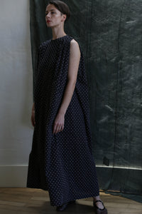 A woman with short hair stands indoors on a wooden floor before a dark green textured backdrop, wearing the Shoulder Pleat Dress in Clip Dot Black & White—an elegant, sleeveless, loose-fitting, ankle-length piece by Atelier Lan Jaenicke.