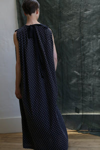 A person with short hair stands with their back to the camera, wearing the Shoulder Pleat Dress | Clip Dot Black & White by Atelier Lan Jaenicke—a long, sleeveless black gown with white dots—against a dark green textured curtain.