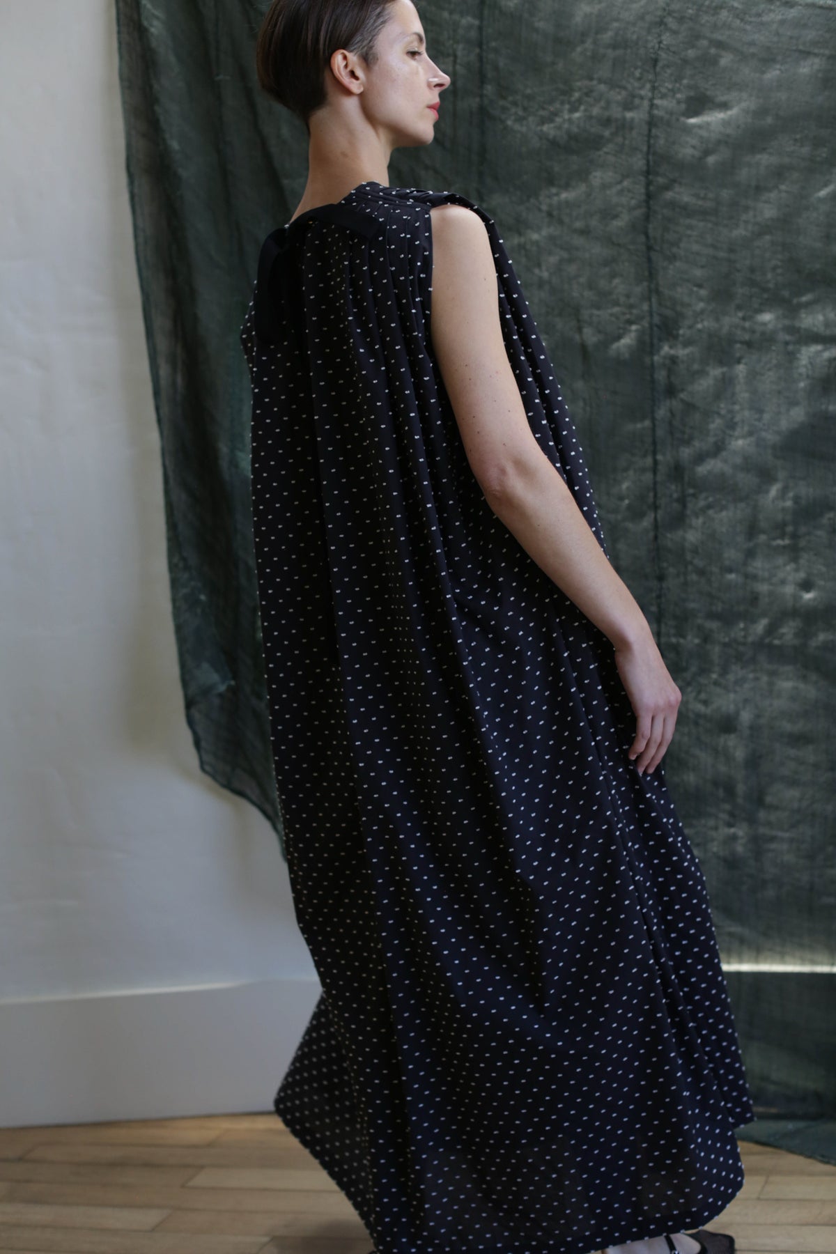 A woman in a long, sleeveless Shoulder Pleat Dress | Clip Dot Black & White stands sideways by a dark green fabric backdrop. With short hair and a relaxed pose, she looks slightly upward, highlighting the Atelier Lan Jaenicke style.