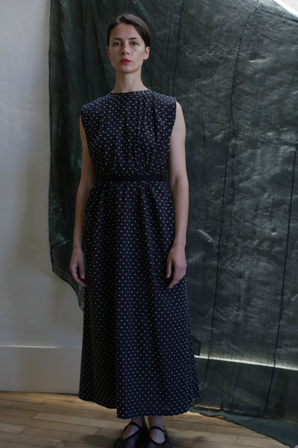 A woman stands indoors wearing the Shoulder Pleat Dress | Clip Dot Black & White—an ankle-length, sleeveless piece with subtle shoulder pleats and white polka dots. Her hair is tied back against a dark green, textured fabric backdrop.