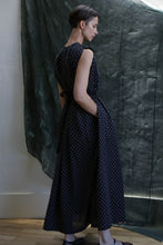Load image into Gallery viewer, A woman in profile wears the Shoulder Pleat Dress | Clip Dot Black &amp; White—sleeveless, ankle-length, with white polka dots and subtle pleats. Her hands rest in her pockets before a dark, textured fabric background.