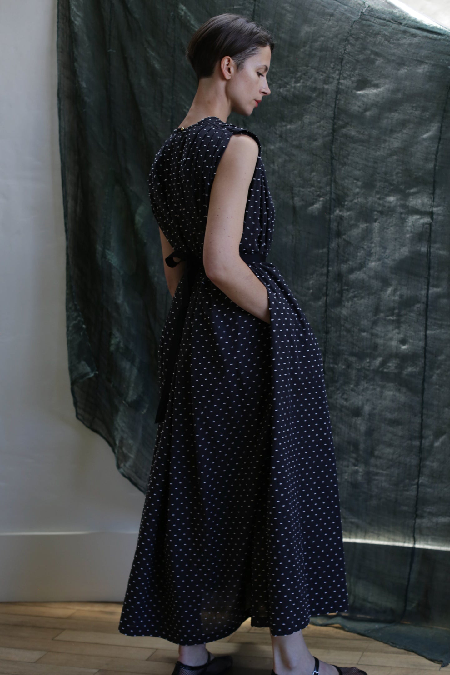 A woman in profile wears the Shoulder Pleat Dress | Clip Dot Black & White—sleeveless, ankle-length, with white polka dots and subtle pleats. Her hands rest in her pockets before a dark, textured fabric background.