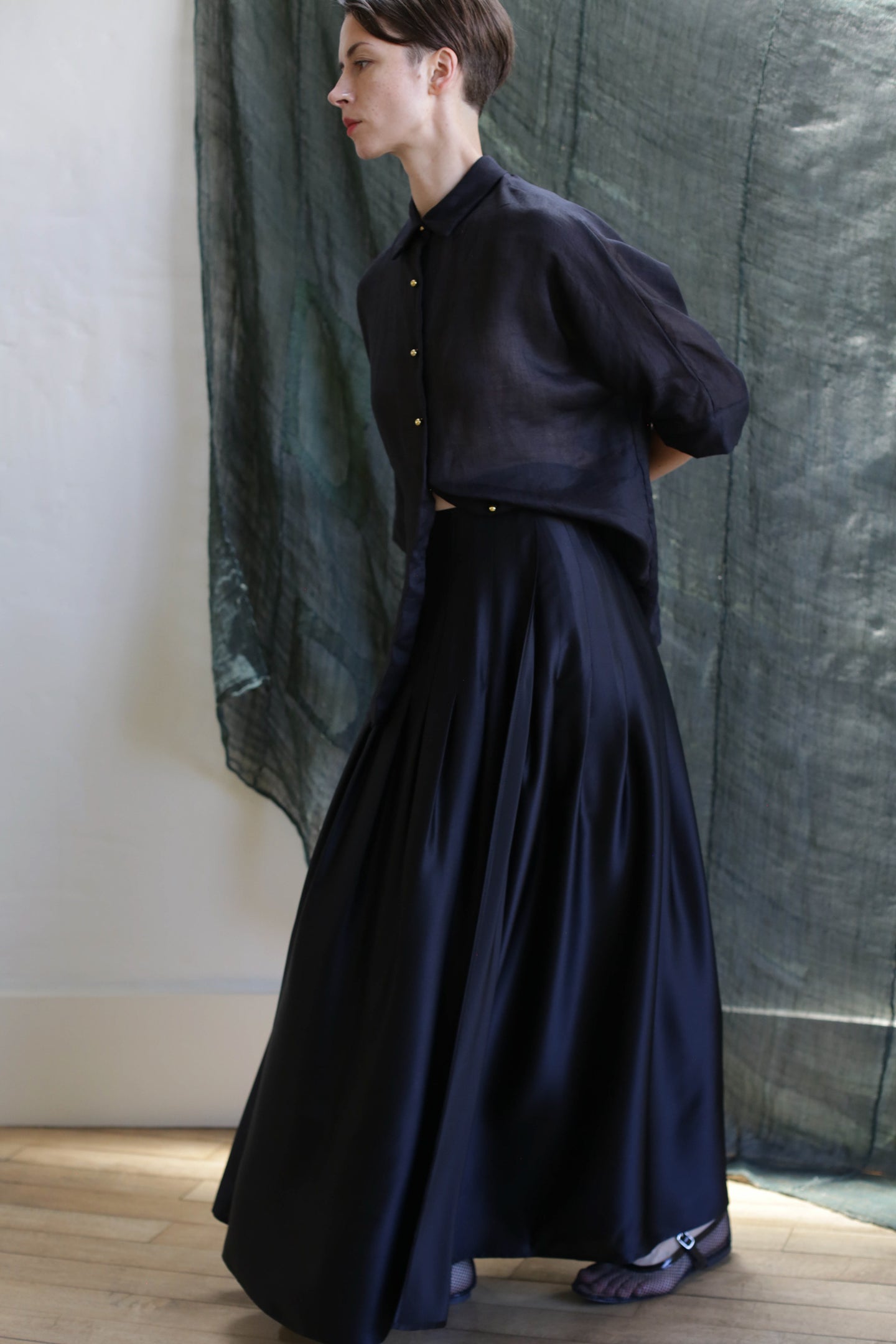 A person in a Hand Pleated Column Wrap Skirt | Silk Charmeuse Black stands in profile against a light wall and textured, semi-transparent greenish curtain, paired with a dark button-up shirt.