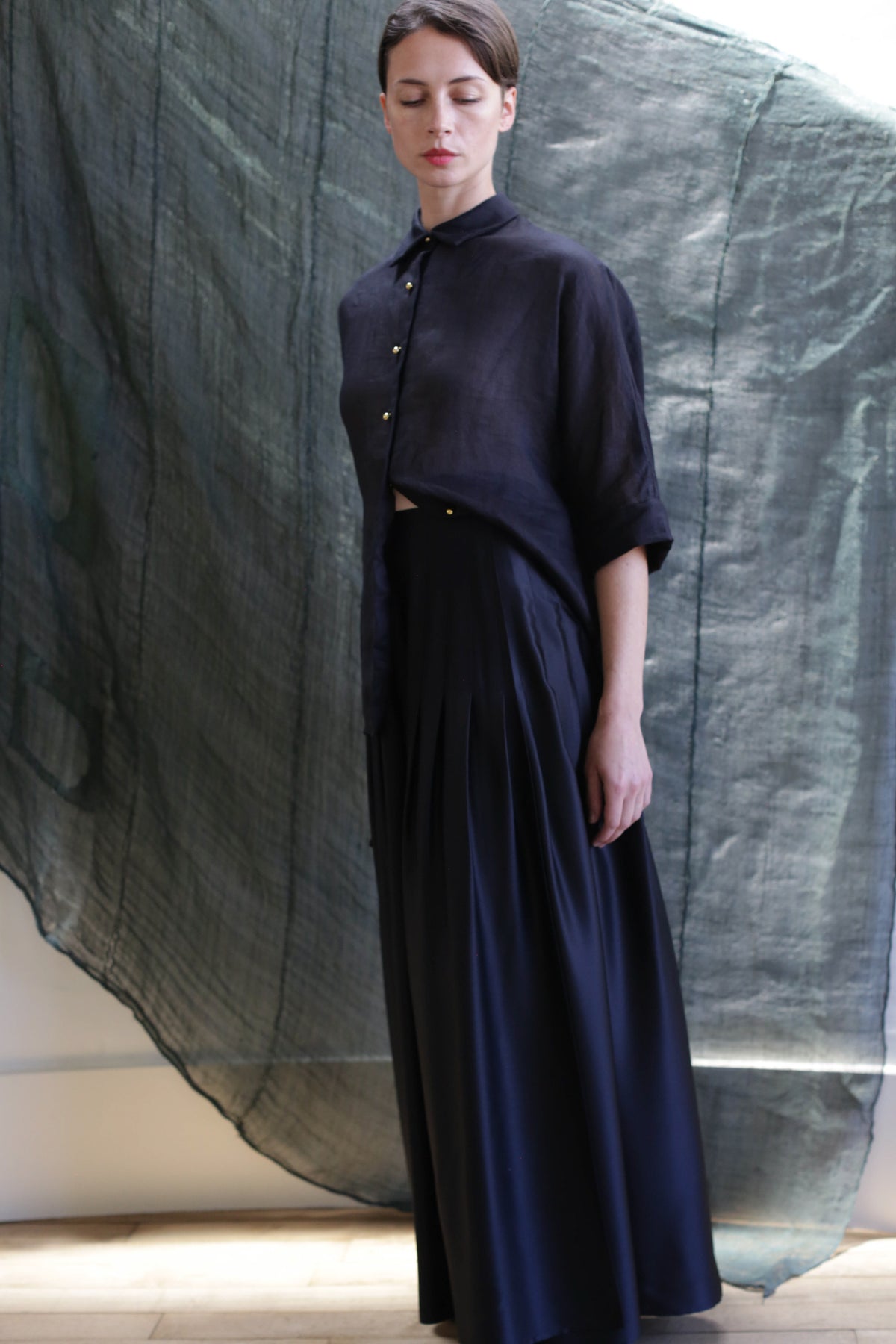 A woman stands before a textured dark green backdrop, wearing a loose button-up shirt and the Hand Pleated Column Wrap Skirt in Black Silk Charmeuse. She gazes down calmly with her hair neatly parted to the side.