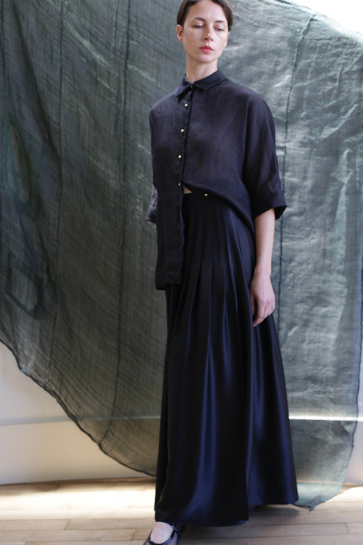 A woman in a loose, dark button-up shirt and the Hand Pleated Column Wrap Skirt | Silk Charmeuse Black stands before a textured dark green backdrop, looking slightly to the side with a calm expression.