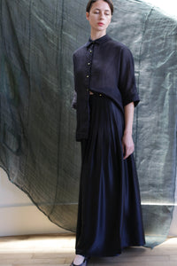 A woman in a loose, dark button-up shirt and the Hand Pleated Column Wrap Skirt | Silk Charmeuse Black stands before a textured dark green backdrop, looking slightly to the side with a calm expression.