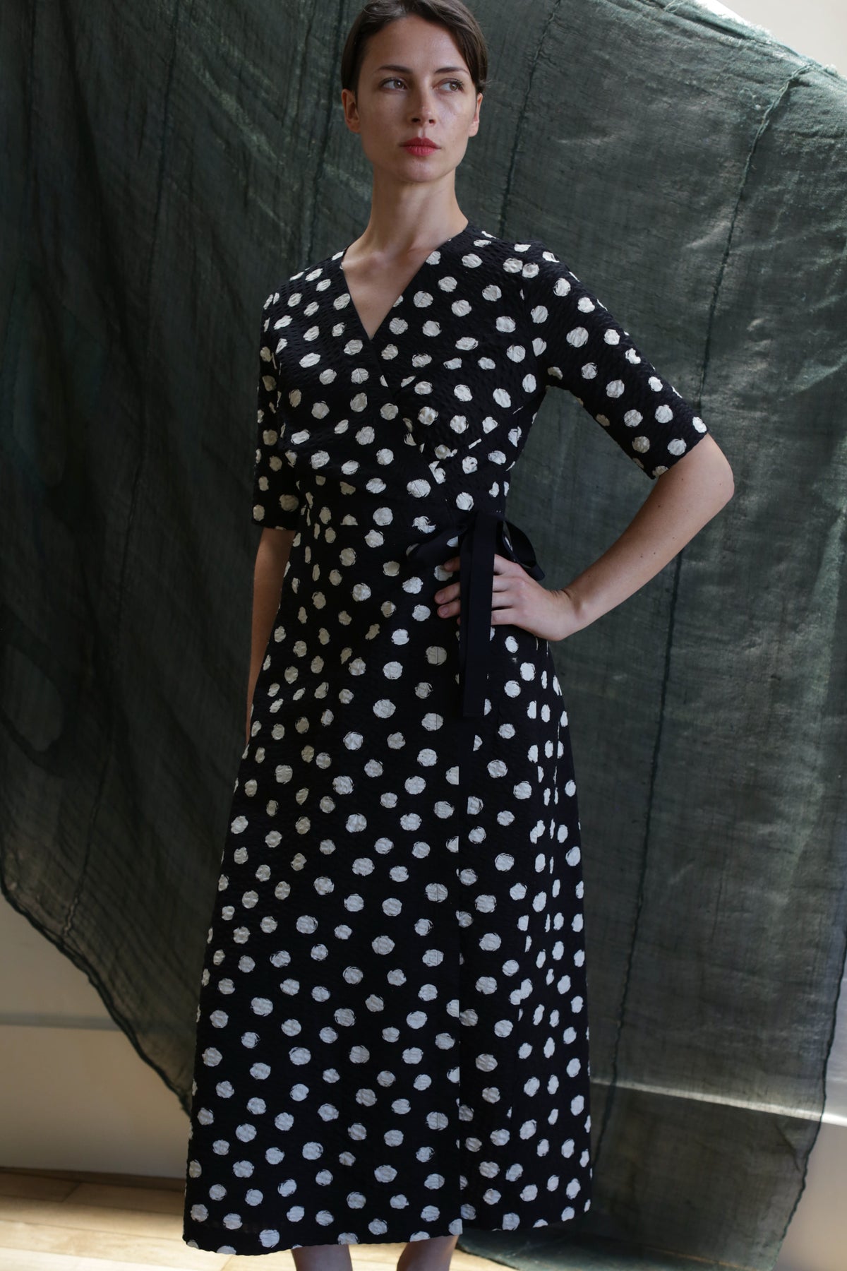 A woman stands with one hand on her hip, wearing the Fitted Wrap Dress in Polkadot Seersucker Black. She looks to the side with a neutral expression, against a backdrop of dark, textured fabric.