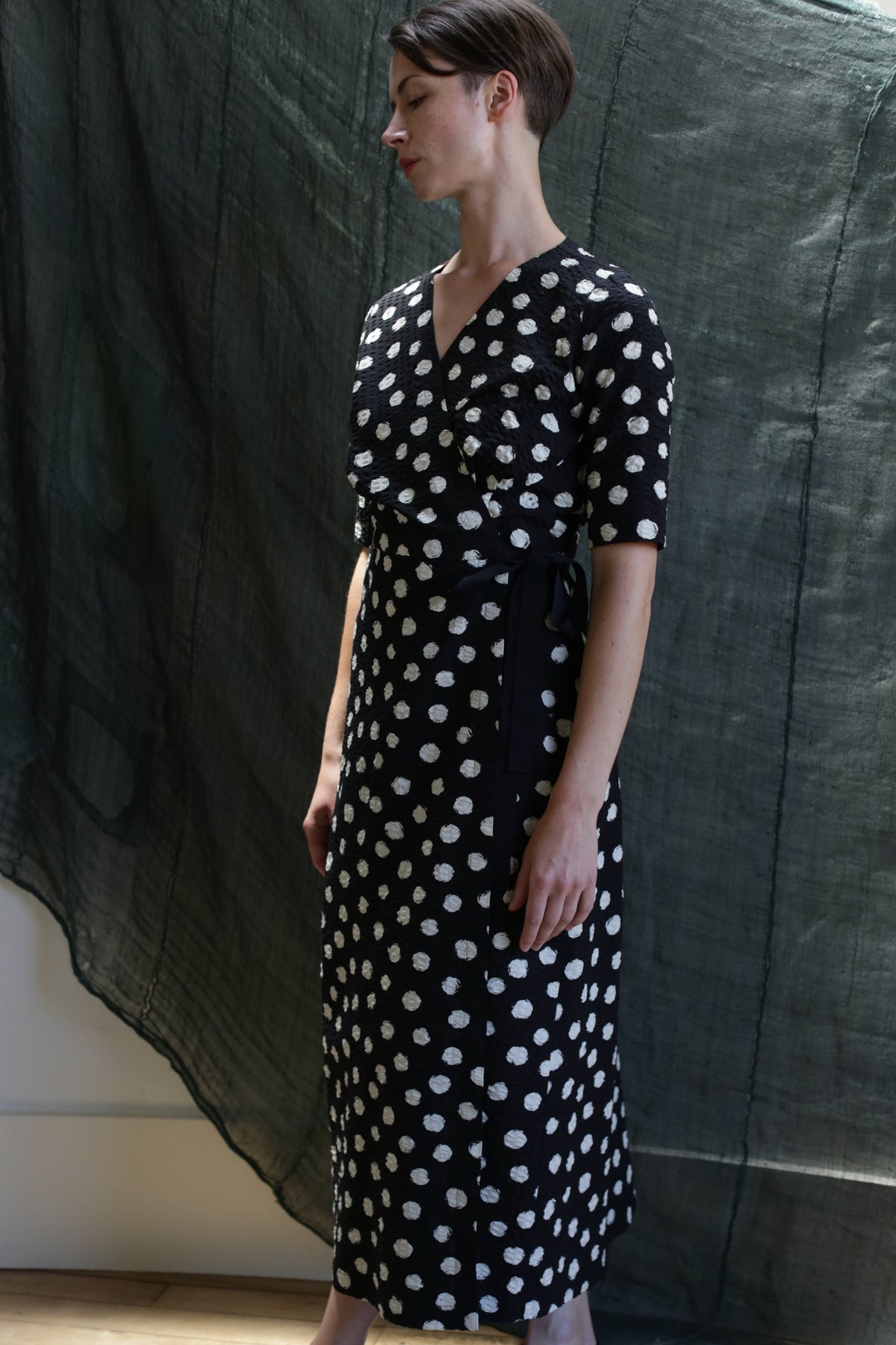 A person stands indoors against a dark, textured fabric backdrop, wearing the Fitted Wrap Dress | Polkadot Seersucker Black, featuring white polka dots, short sleeves, and a side tie, looking slightly downward with hands relaxed by their sides.