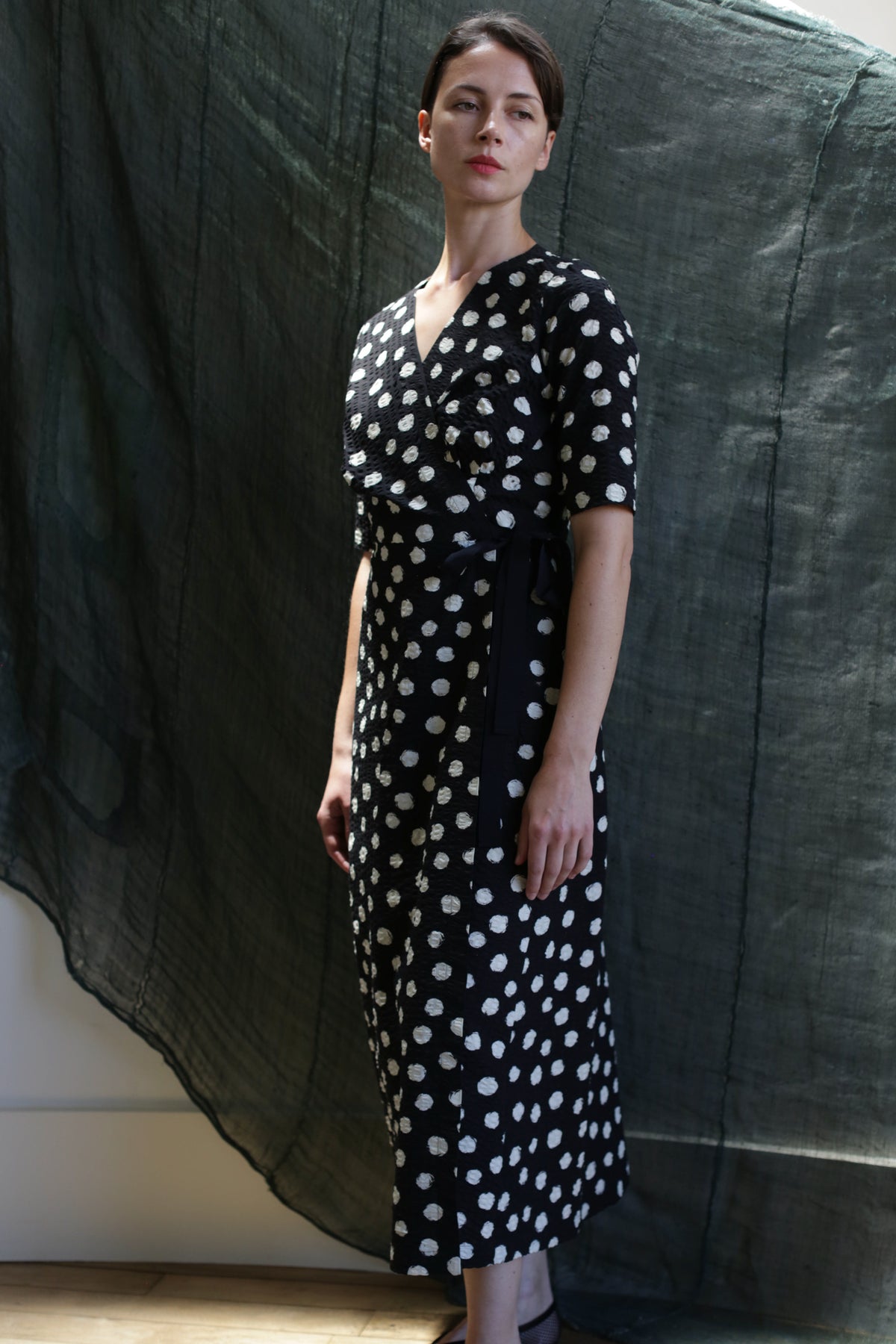 A woman with short dark hair and a neutral expression stands indoors wearing the Fitted Wrap Dress | Polkadot Seersucker Black, a black calf-length dress with white polka dots and a sash belt, set against a backdrop of dark textured fabric.