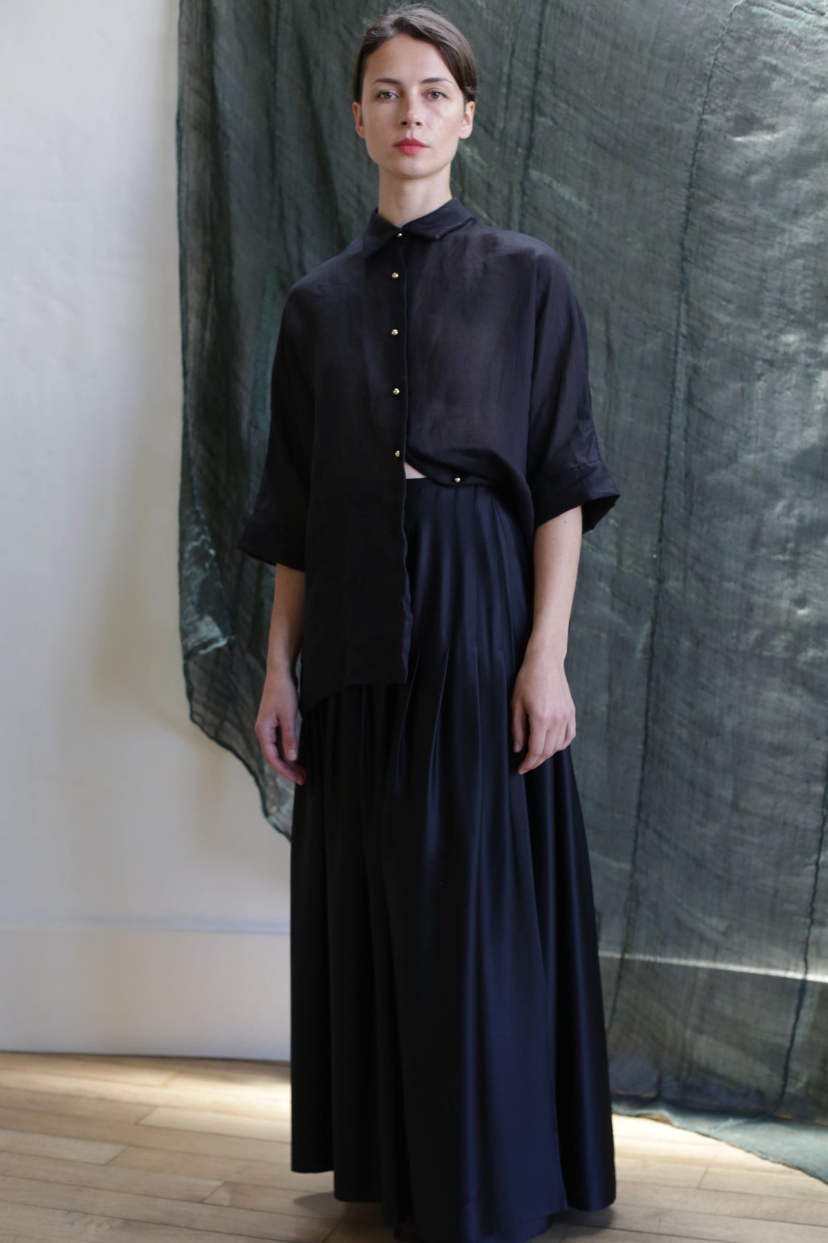 A person wears the Hand Pleated Column Wrap Skirt in Silk Charmeuse Black, paired with a loose black button-up top, standing indoors against a dark green fabric backdrop with soft natural light and wooden floors.
