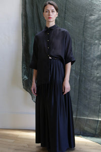 A person wears the Hand Pleated Column Wrap Skirt in Silk Charmeuse Black, paired with a loose black button-up top, standing indoors against a dark green fabric backdrop with soft natural light and wooden floors.