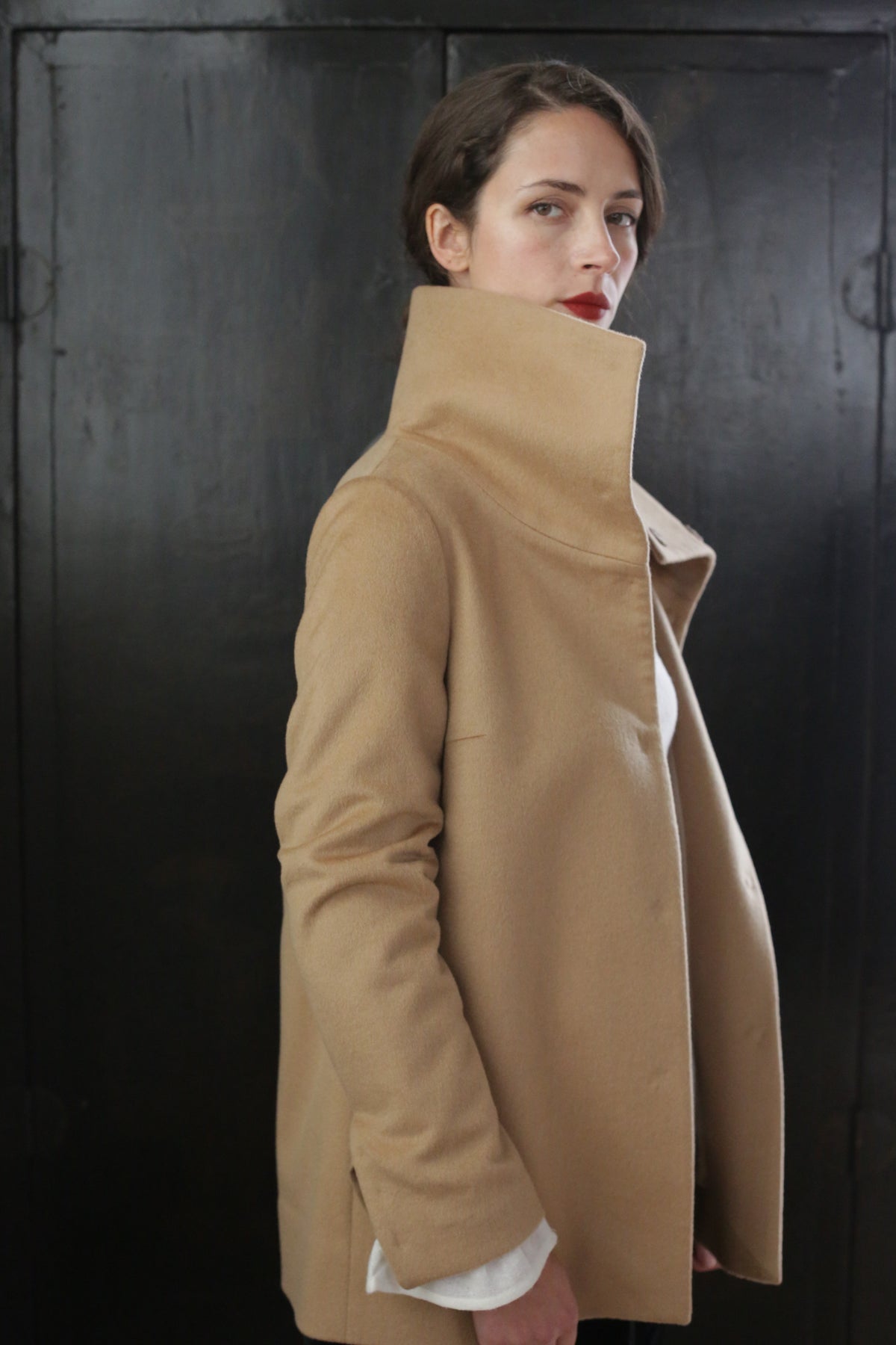 A woman with short dark hair stands in front of a dark background, wearing the Stamford II | Camel made from 100% woven cashmere. Her expression is neutral, and she gazes slightly to the side, with her right arm bent and her left arm at her side.