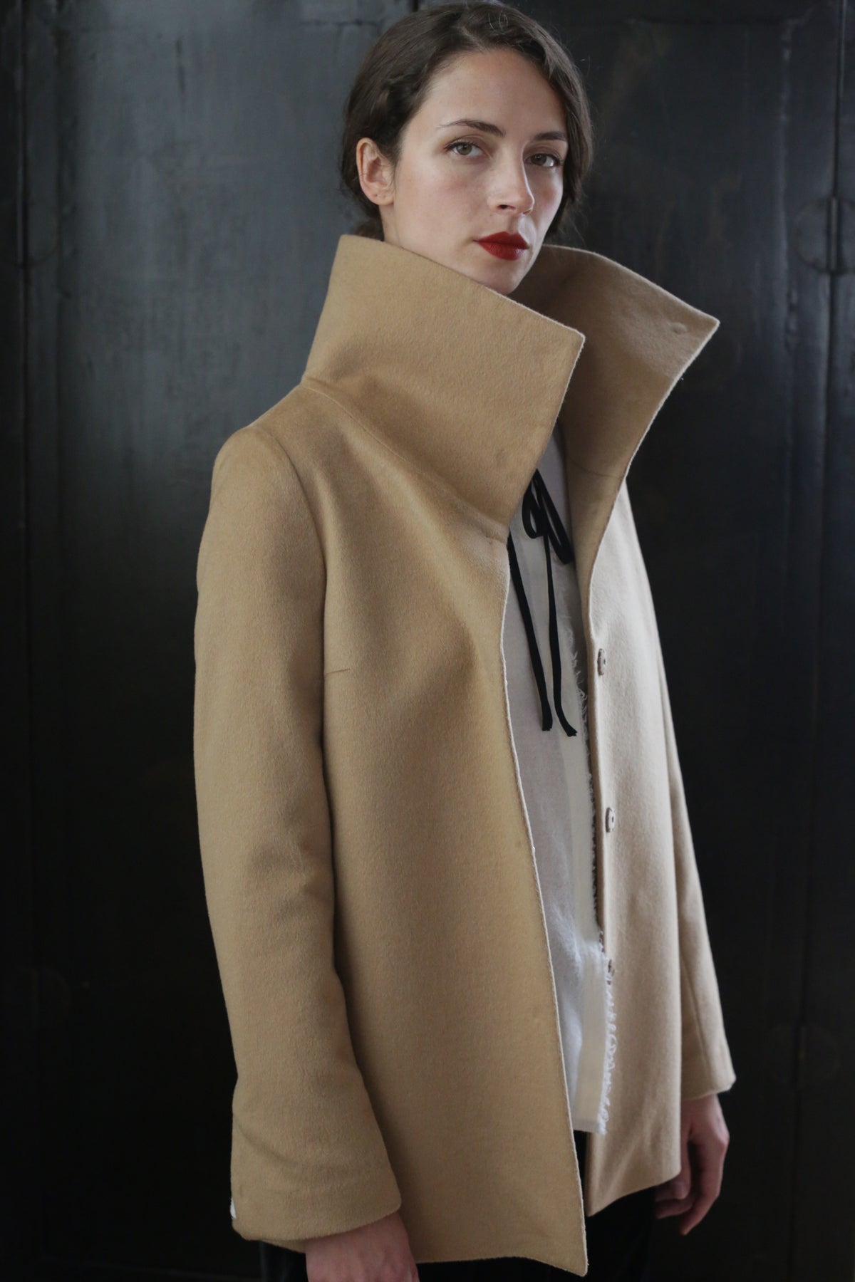 A person with short dark hair stands in front of a dark background, wearing the Stamford II | Camel over a white shirt. The signature jacket, crafted from 100% woven cashmere, drapes perfectly as the individual's head is slightly tilted, exuding an air of confidence and poise.