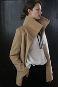 A person with their hair tied back is standing sideways, wearing a signature beige coat with a high collar over a white blouse. Their left hand is in the coat pocket, and the background is a dark, textured surface. This Stamford II | Camel jacket is crafted from 100% woven cashmere for unparalleled elegance.