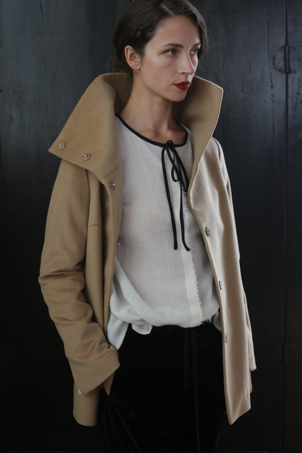 A person with short hair, wearing a Stamford II | Camel made of 100% woven cashmere over a white blouse with a black tie at the collar, stands against a dark background. The individual's expression is serious, and they have one hand in their pocket.