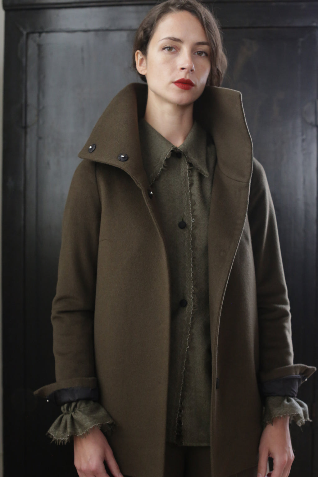 A woman with short brown hair stands against a dark background. She is wearing her signature jacket—a Stamford II | Olive with a high collar over a green top. The rich cashmere fabric adds an air of sophistication as she gazes directly at the camera with a neutral expression.