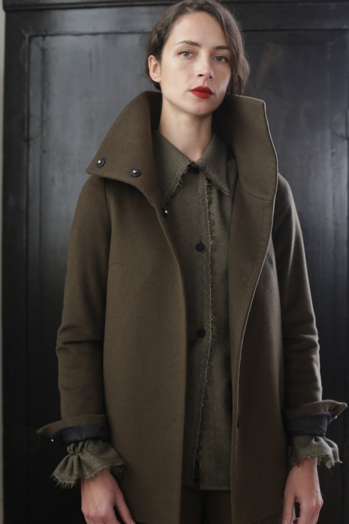 A woman with short brown hair stands against a dark background. She is wearing her signature jacket—a Stamford II | Olive with a high collar over a green top. The rich cashmere fabric adds an air of sophistication as she gazes directly at the camera with a neutral expression.