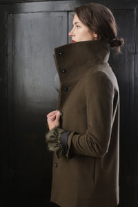 A woman in an olive green cashmere coat stands in profile with the collar turned up. She has her dark hair tied in a low bun and holds the coat with her left hand. The background is dark and features a textured surface, highlighting the elegance of this Stamford II | Olive jacket.