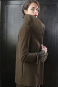 A person with dark hair styled in an updo, wearing a high-collared Stamford II | Olive coat, stands against a dark background. They glance over their shoulder with a serious expression. The coat's cuff features a fur-like embellishment.