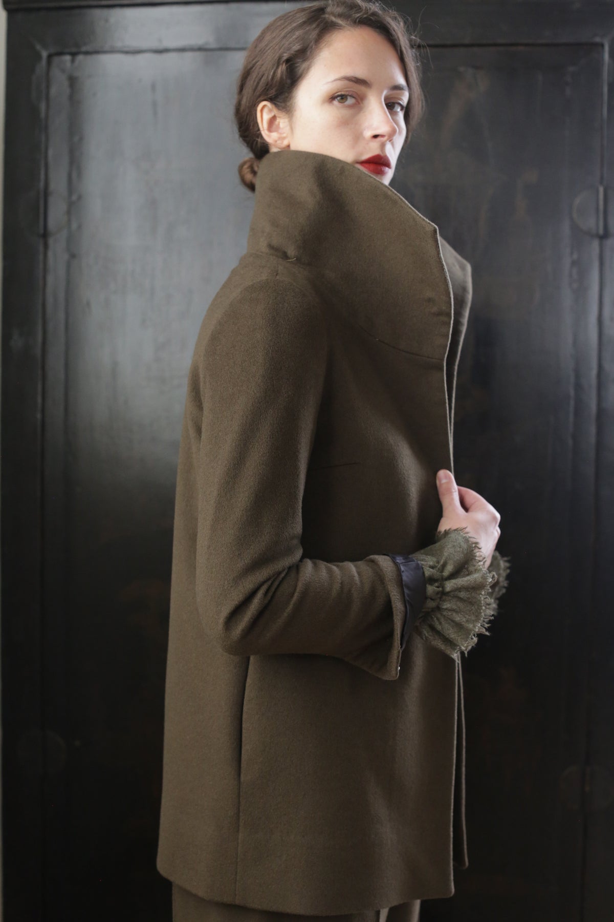 A person with dark hair styled in an updo, wearing a high-collared Stamford II | Olive coat, stands against a dark background. They glance over their shoulder with a serious expression. The coat's cuff features a fur-like embellishment.