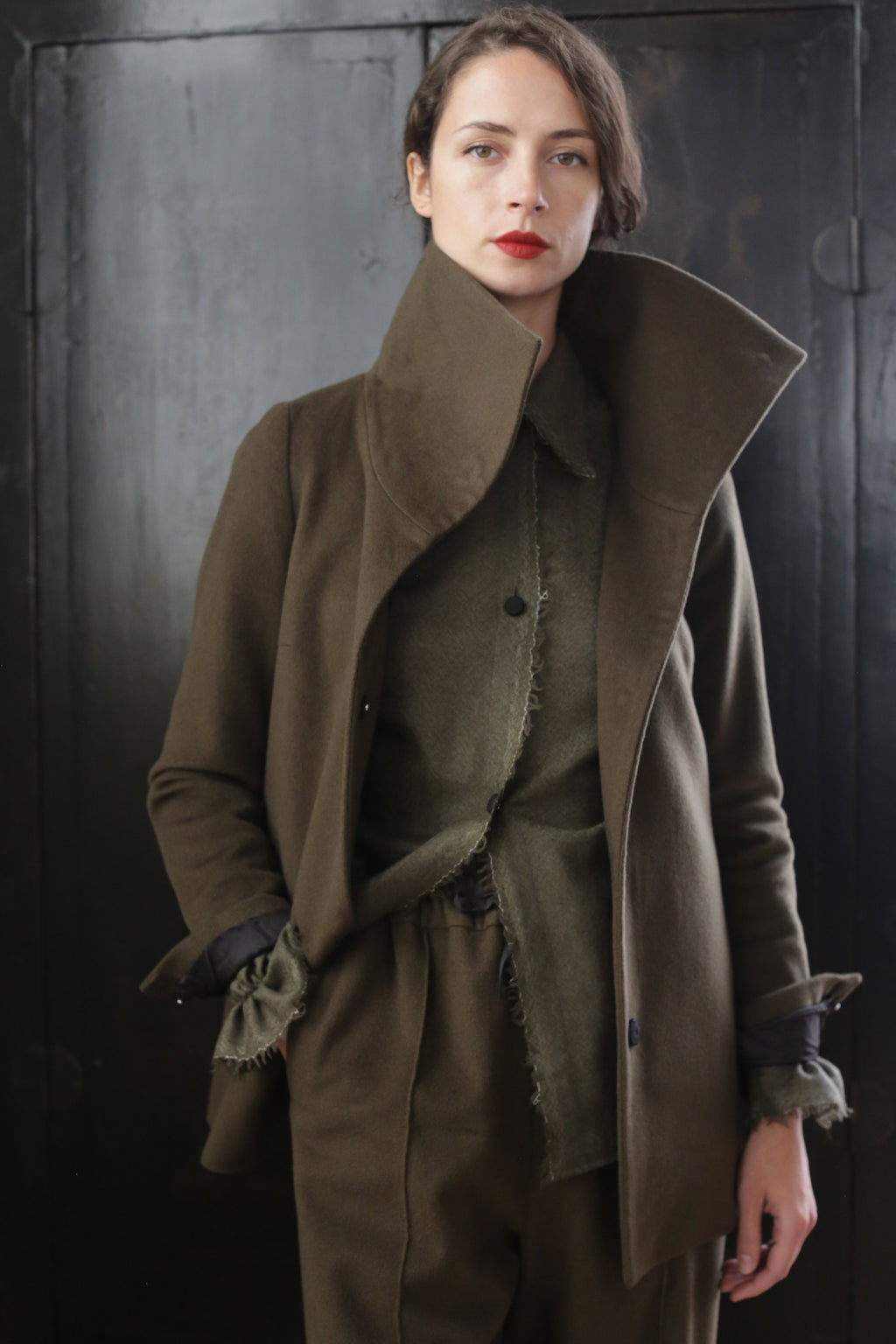 A person stands against a dark background, wearing a stylish Stamford II | Olive with a high collar, and matching pants. Their hair is short, and they have a serious expression while looking directly at the camera.