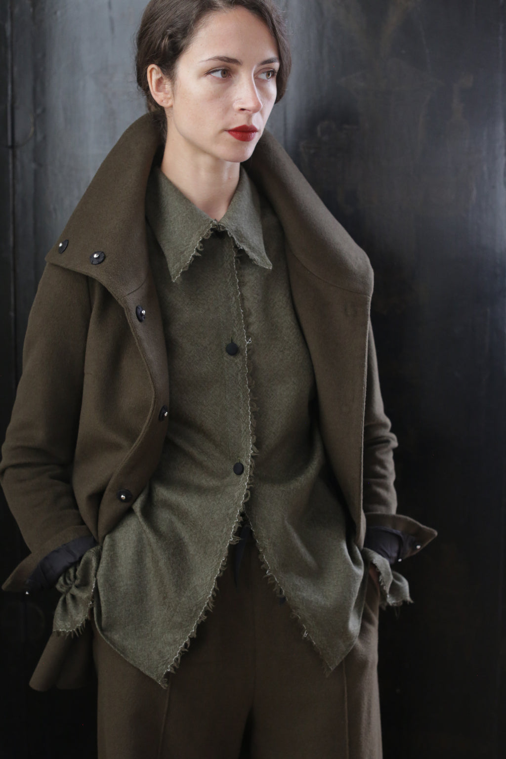 A person wearing a stylish, dark olive green Stamford II | Olive jacket over a matching top and pants stands with hands in their pockets. They have short, dark hair, a neutral expression, and are positioned against a dark, glossy background.