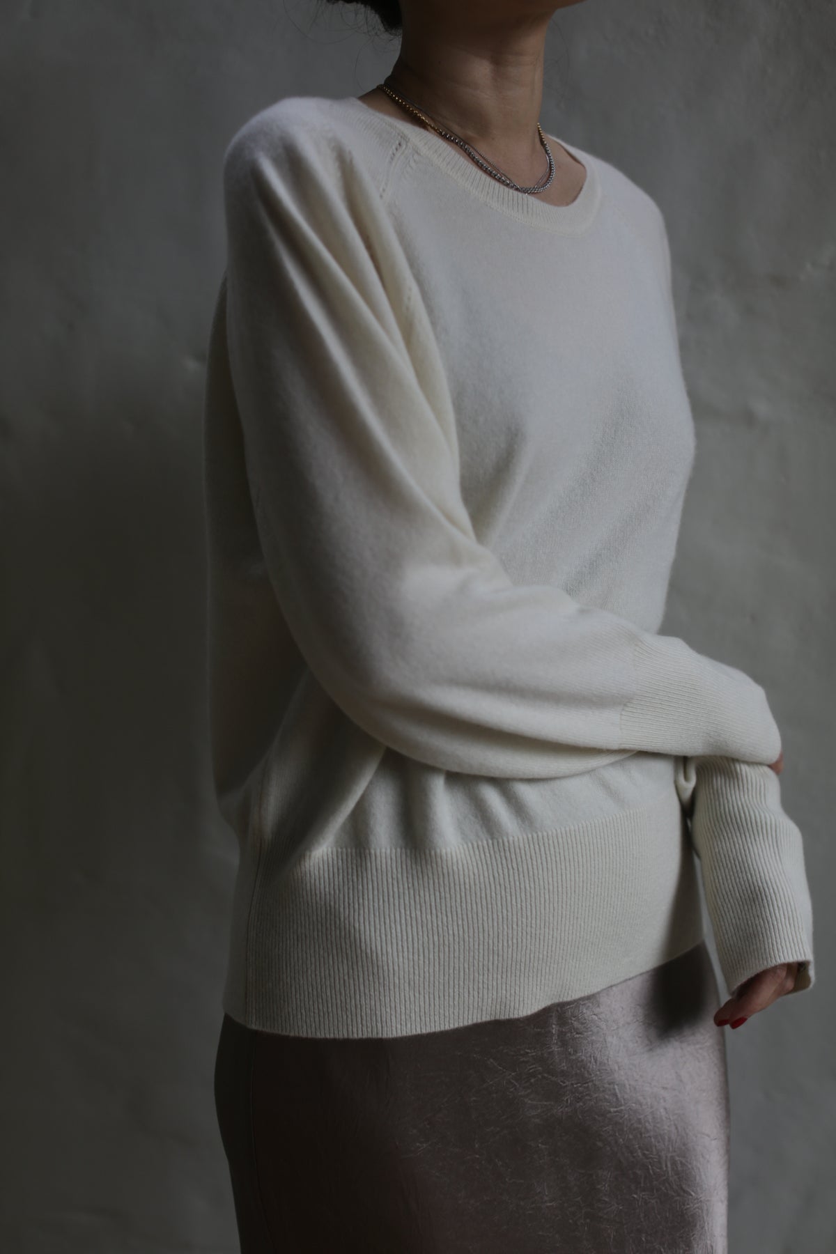 A person stands against a textured wall wearing a Raglan Crewneck Cashmere Sweater | Ivory and a metallic skirt. The person is partially cropped out of the frame, with their head not visible. The light source creates a soft, shadowy effect on the clothing.