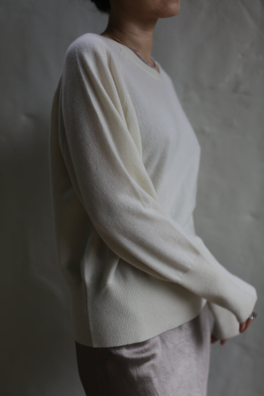 A person is shown from the neck down wearing a light, long-sleeved Raglan Crewneck Cashmere Sweater | Ivory and a skirt. The background is plain and indistinct. The person's right arm is slightly bent and their left arm hangs down naturally. The lighting is soft, creating a gentle shadow.