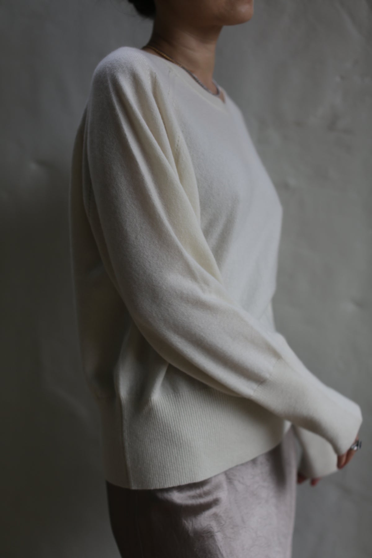 A person is shown from the neck down wearing a light, long-sleeved Raglan Crewneck Cashmere Sweater | Ivory and a skirt. The background is plain and indistinct. The person's right arm is slightly bent and their left arm hangs down naturally. The lighting is soft, creating a gentle shadow.