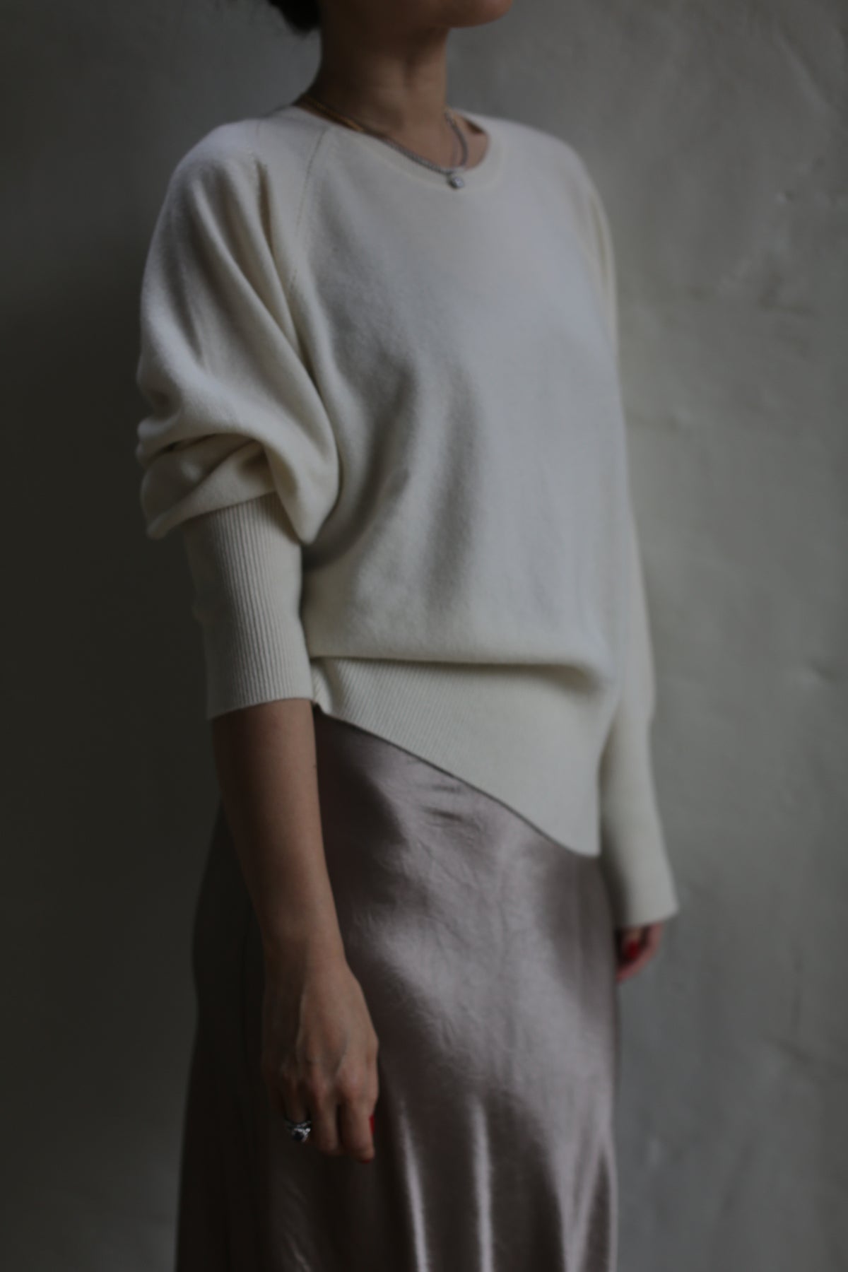 A person dressed in a white, long-sleeved Raglan Crewneck Cashmere Sweater | Ivory and a light, silky skirt stands against a plain, textured gray wall. The image captures the outfit from the shoulders down, with the head slightly cropped out of the frame.