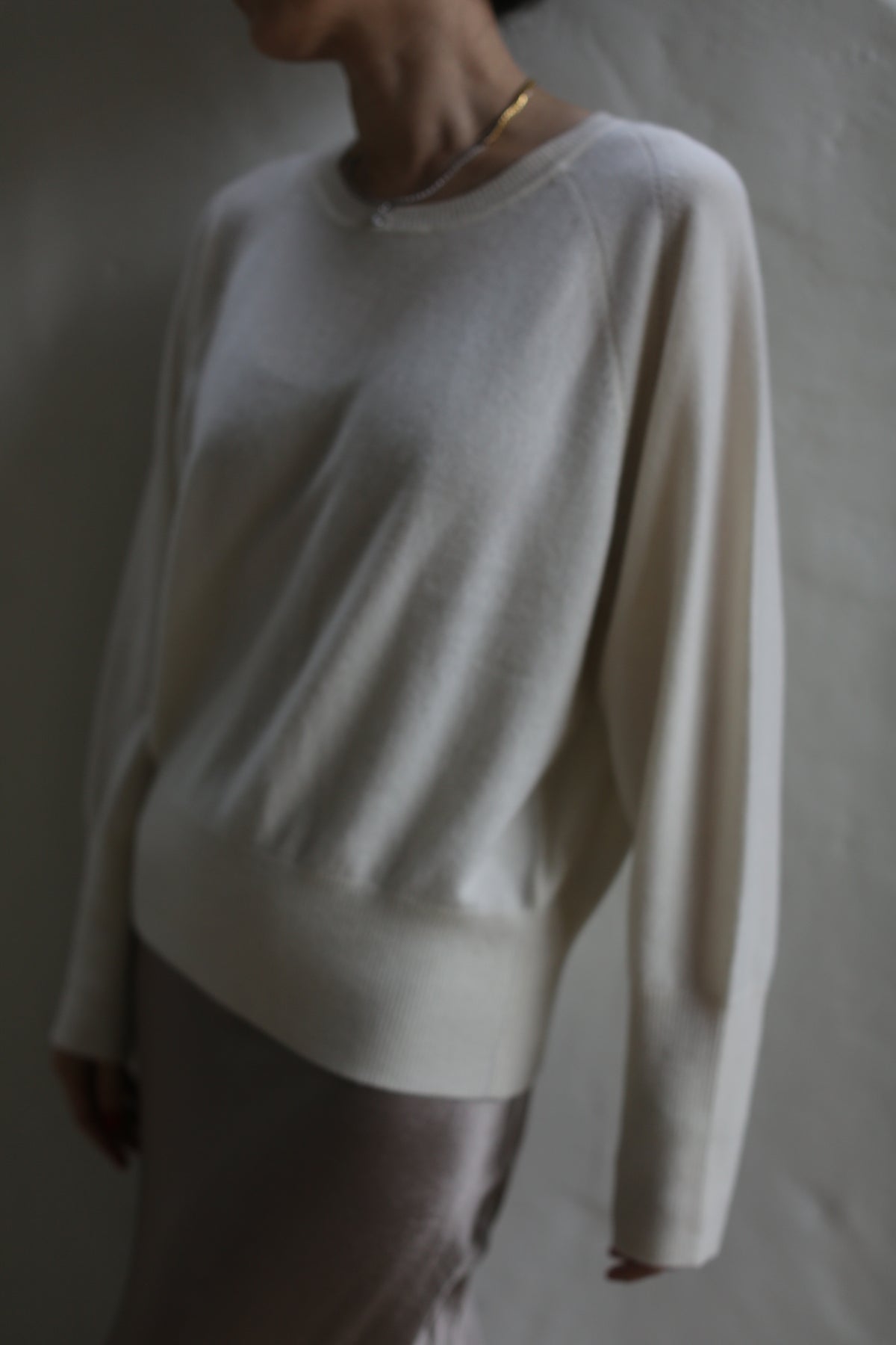 A person wearing a light, long-sleeved Raglan Crewneck Cashmere Sweater | Ivory and a silky, light-colored skirt stands against a wall. The image is slightly out of focus, creating a soft, blurry effect. The person's head is partially visible and slightly turned to the side.