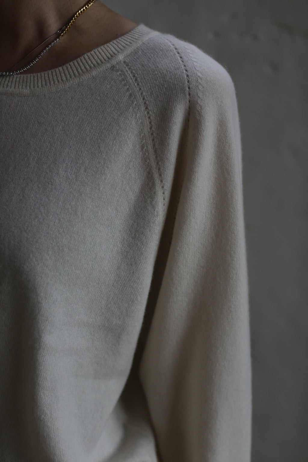 A close-up of a person wearing a light-colored, long-sleeved, Raglan Crewneck Cashmere Sweater | Ivory with raglan sleeves and a delicate gold necklace. The focus is on the shoulder area, with the background blurred, emphasizing the texture and softness of the fabric.