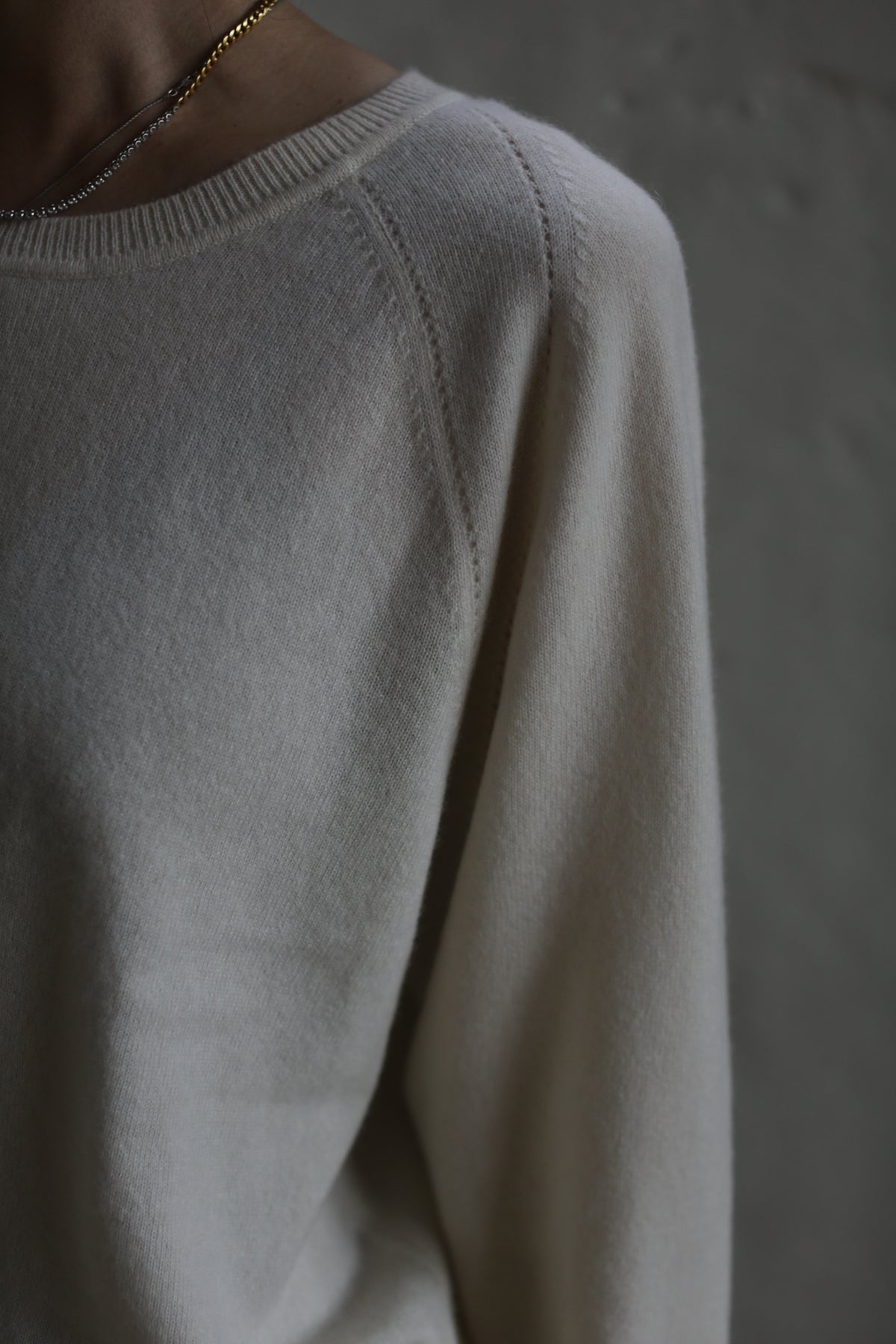 A close-up of a person wearing a light-colored, long-sleeved, Raglan Crewneck Cashmere Sweater | Ivory with raglan sleeves and a delicate gold necklace. The focus is on the shoulder area, with the background blurred, emphasizing the texture and softness of the fabric.