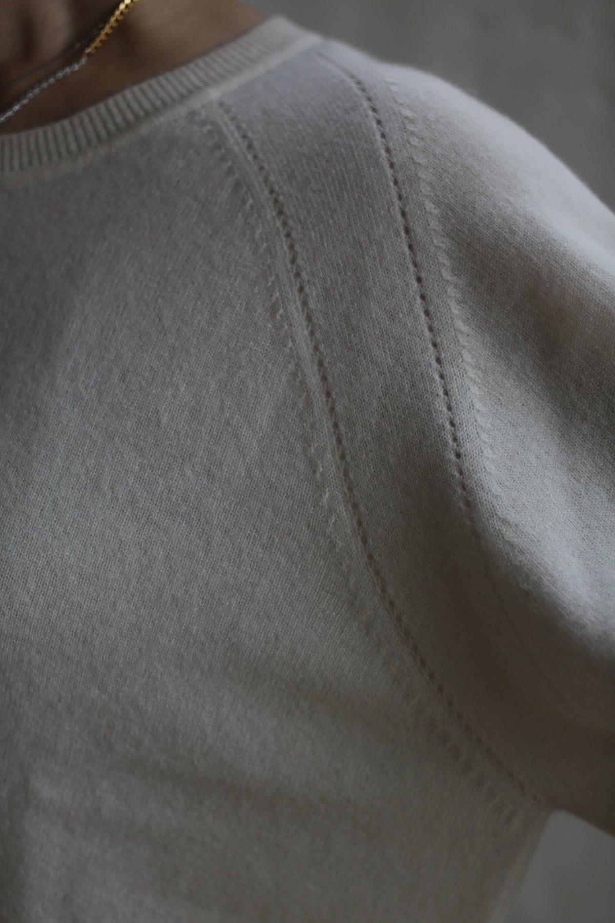Close-up of a person wearing a Raglan Crewneck Cashmere Sweater | Ivory. The focus is on the shoulder and upper sleeve, highlighting the stitching and texture of the fabric. The edge of a gold necklace is faintly visible at the crewneck. The background is softly blurred.