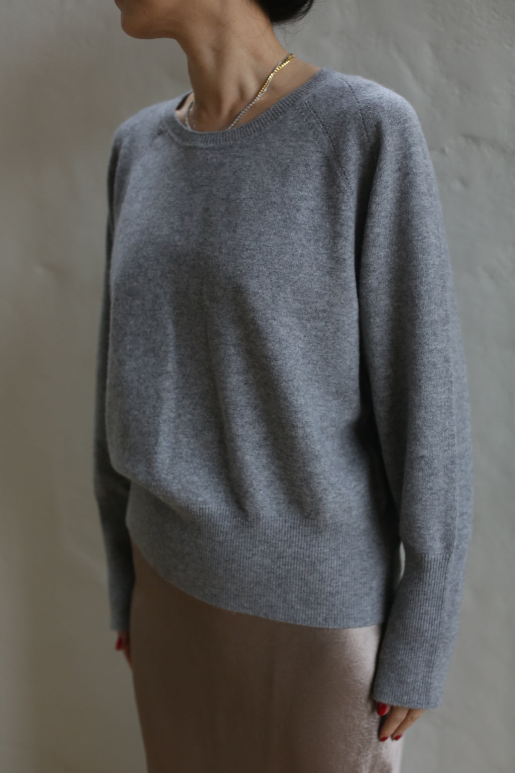 A person wearing a Raglan Crewneck Cashmere Sweater | Grey and a beige skirt stands against a neutral wall. They have a gold necklace, and their face is not visible as they are turned sideways. This crewneck sweater is truly a wardrobe staple.