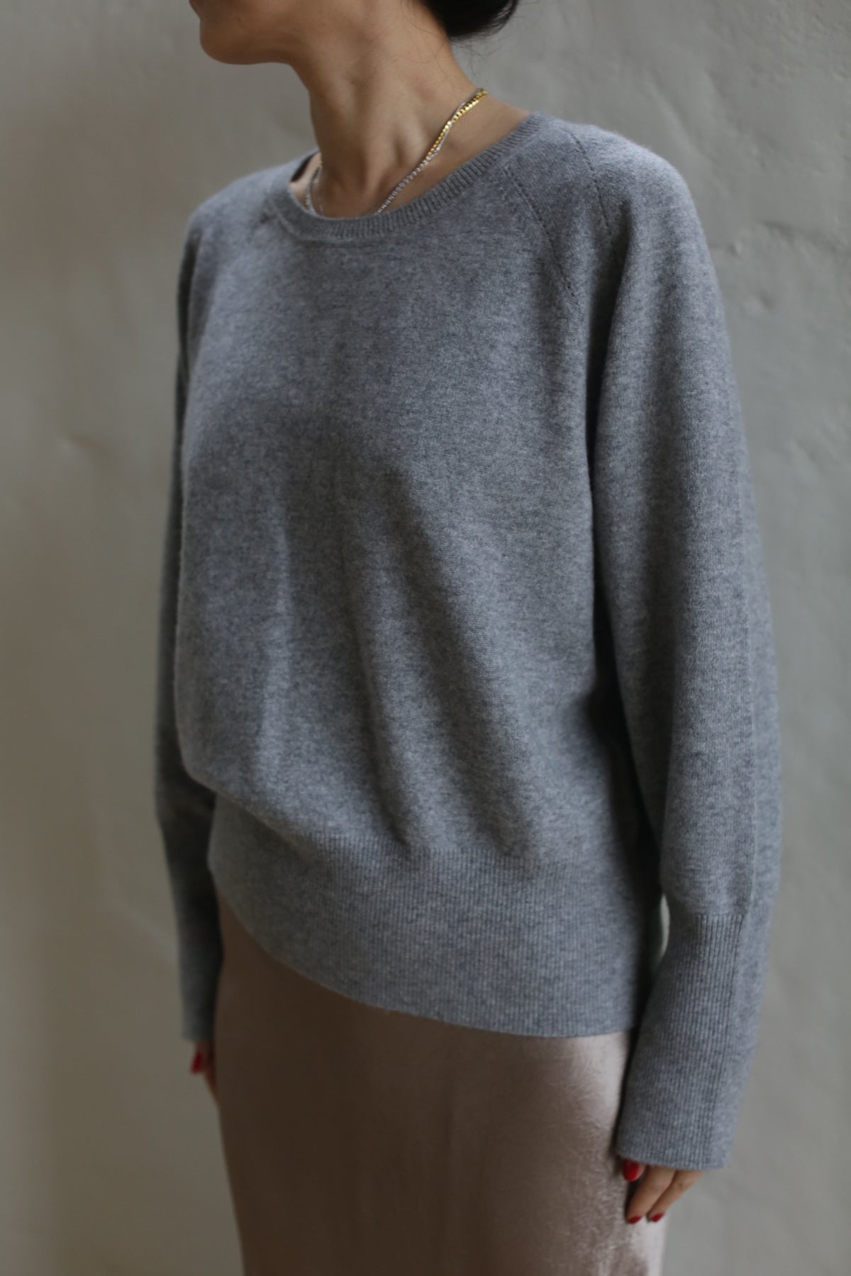 A person wearing a Raglan Crewneck Cashmere Sweater | Grey and a beige skirt stands against a neutral wall. They have a gold necklace, and their face is not visible as they are turned sideways. This crewneck sweater is truly a wardrobe staple.