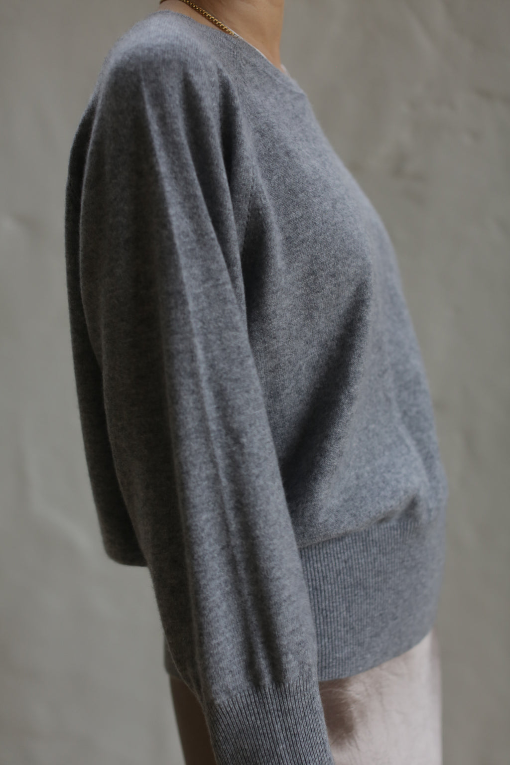 A person is wearing a grey, long-sleeved Raglan Crewneck Cashmere Sweater. The image is focused on the upper torso, showcasing the texture and relaxed fit of this wardrobe staple. The background is a plain, light-colored wall. The person's face is not visible.