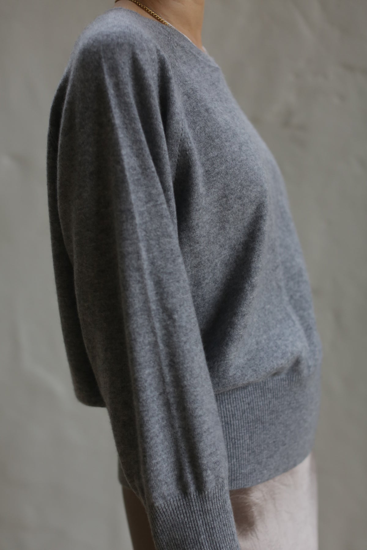 A person is wearing a grey, long-sleeved Raglan Crewneck Cashmere Sweater. The image is focused on the upper torso, showcasing the texture and relaxed fit of this wardrobe staple. The background is a plain, light-colored wall. The person's face is not visible.