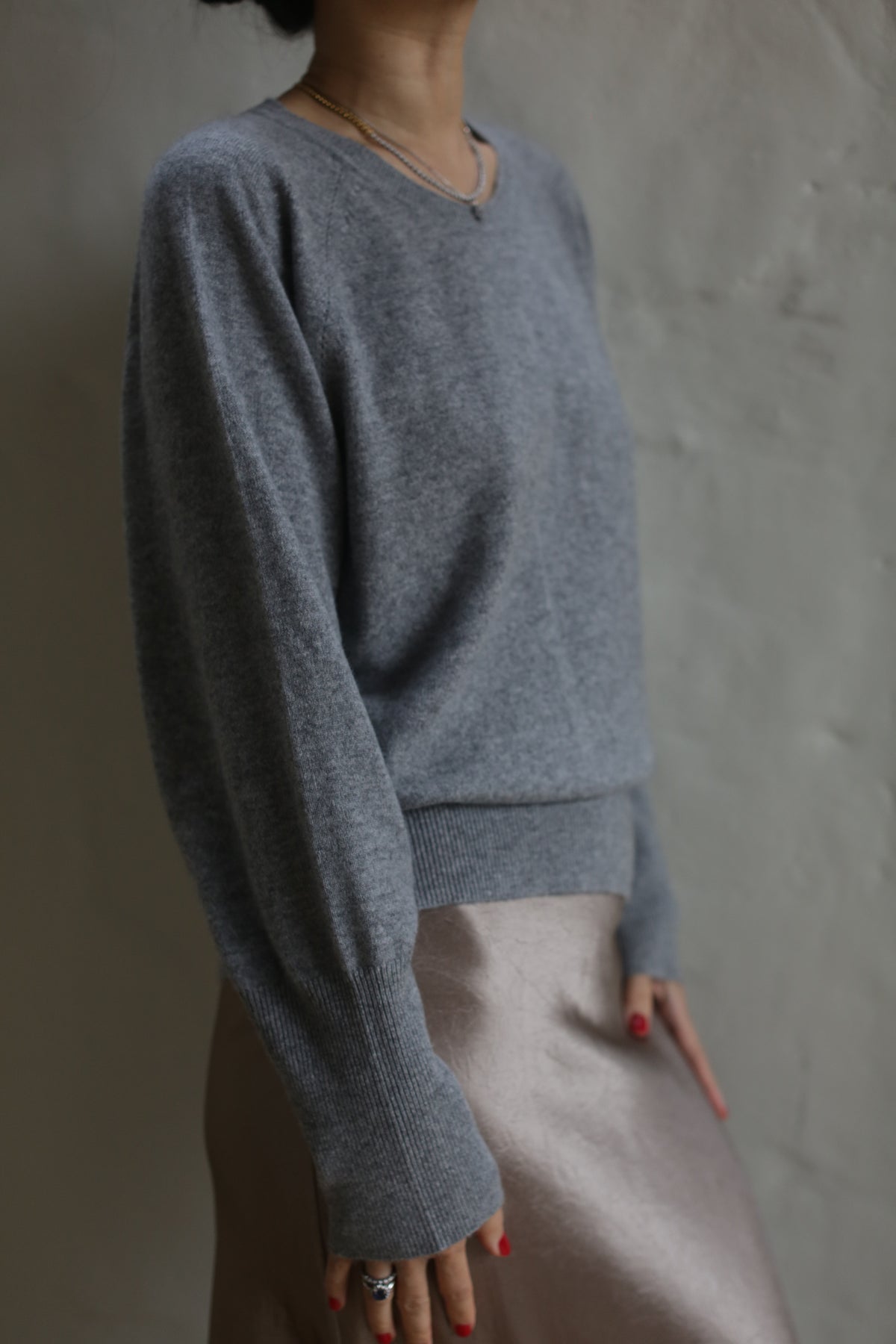 A person wearing a loose-fitting Raglan Crewneck Cashmere Sweater | Grey and a metallic skirt stands in front of a neutral-colored wall. The person’s face is not visible as their head is slightly turned up and away from the camera. The photo has soft, moody lighting, highlighting the sweater as a timeless wardrobe staple.