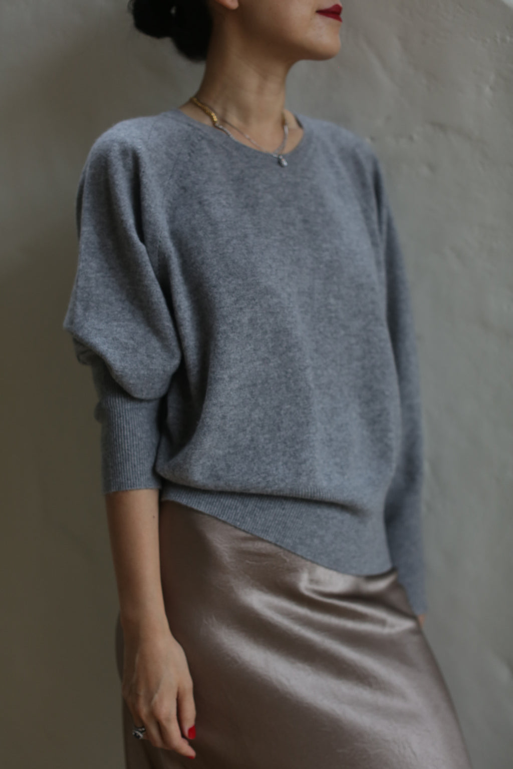 A person wearing a Raglan Crewneck Cashmere Sweater | Grey and a shiny taupe skirt stands against a plain wall. The individual has short hair and wears a necklace. Their face is not fully visible, and their hands rest at their sides.