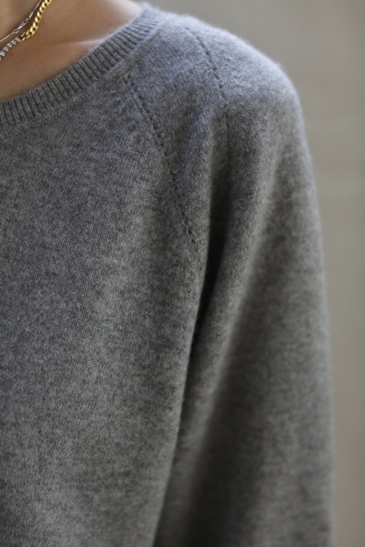 Close-up of a person's upper body wearing a Raglan Crewneck Cashmere Sweater | Grey. The image focuses on the texture and stitching details of the fabric, highlighting this wardrobe staple. The person's neck and part of a gold necklace are slightly visible at the top left corner.