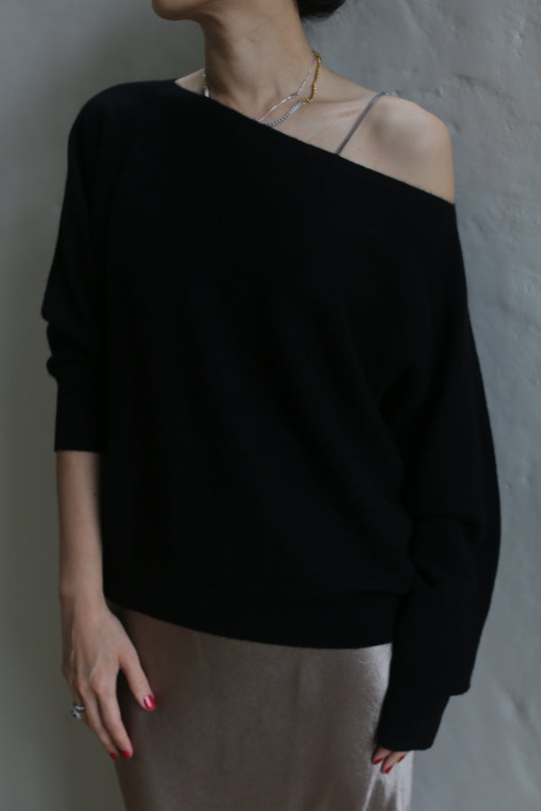 A person dons the Ballerina's Cashmere Sweater in black, revealing a gray strap underneath. With their head slightly tilted, they stand against a plain, light-colored background. One hand hangs by their side, and only the upper part of their body is visible.